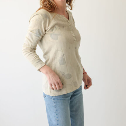 A woman wears a grey/cream silk cotton and cashmere long sleeved jersey top with V neck and buttons half way down the front. It has been printed with lilac cotinus leaf eco prints Side view