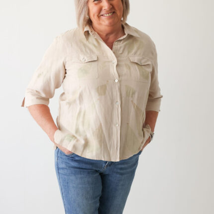 A happy woman wears a light beige silk and linen mix safari suit style short sleeve shirt with two breast pockets. The shirt has been eco printed with large individual green peony leaf prints