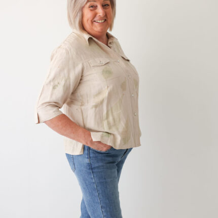 A happy woman wears a light beige silk and linen mix safari suit style short sleeve shirt with two breast pockets. The shirt has been eco printed with large individual green peony leaf prints