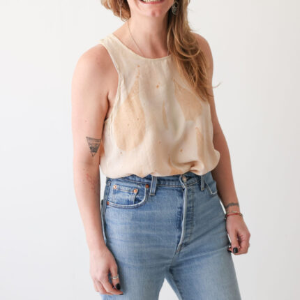 A smiling woman wearing pale blue jeans has paired them with a peachy cream silk vest top eco printed with large brown horse chestnut leaf prints