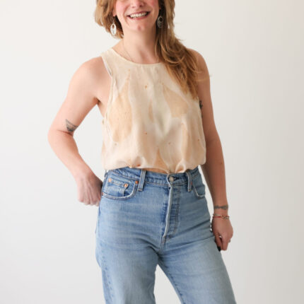 A smiling woman wearing pale blue jeans has paired them with a peachy cream silk vest top eco printed with large brown horse chestnut leaf prints